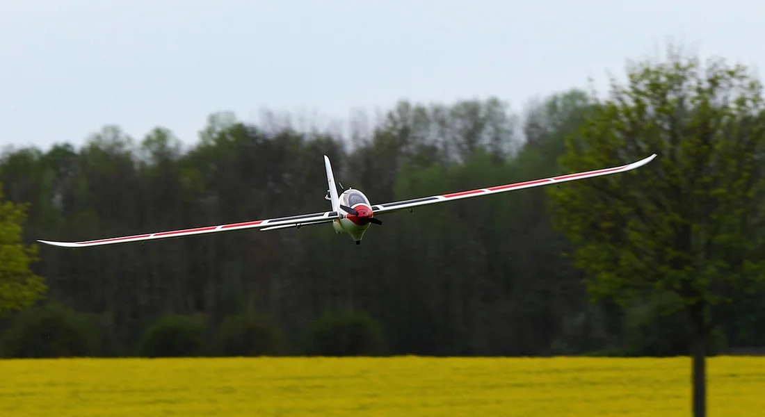 A small glider-style airplane flies toward the camera over a bright yellow field with a tree line in the distance.