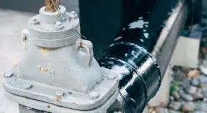 Outdoor close-up of a metal backflow prevention valve attached to a dark pipe on an exterior setting.