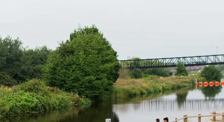 Riverside scene with trees and shrubs along the water, a pedestrian path, orange safety barriers on the bank, and a distant metal bridge.