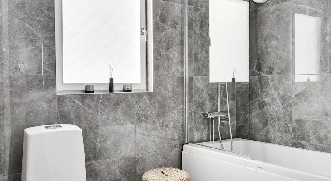 Modern bathroom showing a white toilet beside a bathtub with gray tile walls, a small stool, and a window.