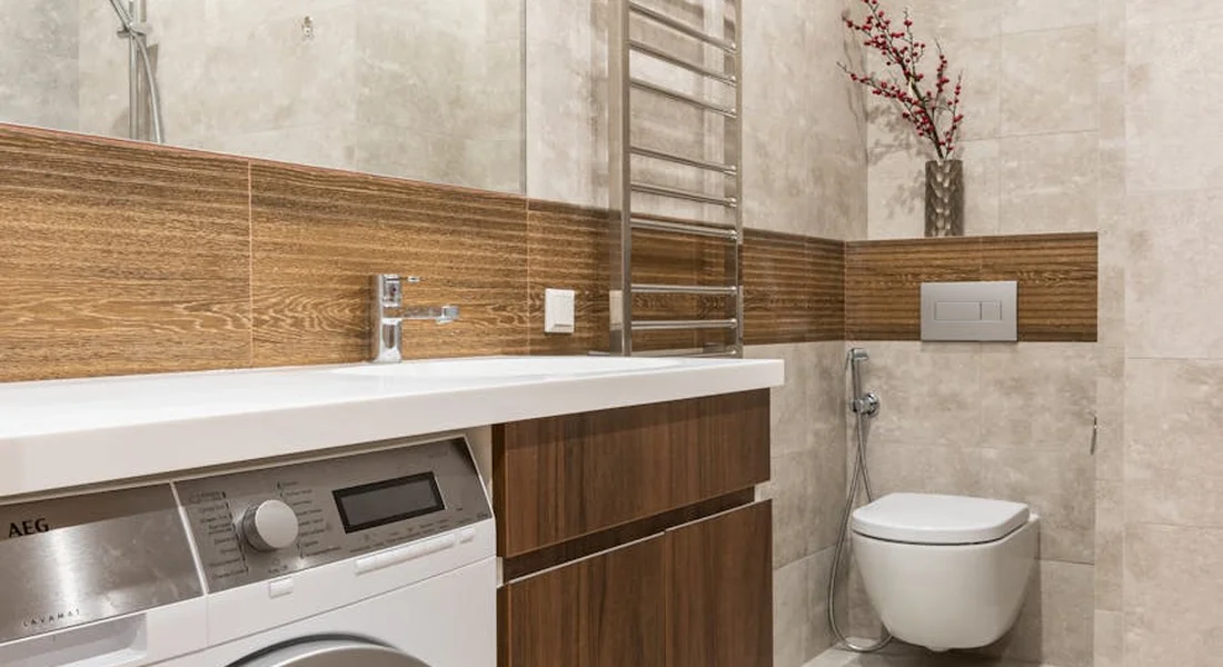 Modern bathroom with a washing machine tucked under a white countertop and wooden vanity, a wall-mounted toilet with a flush plate, a towel rack, and a decorative vase on a shelf.