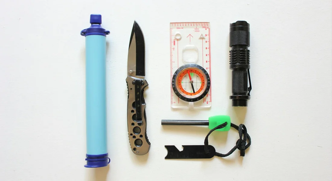 Flat lay of camping gear for water purification, including a blue portable water filter, a folding knife, a compass, a ruler, a small flashlight, and a keyring whistle.