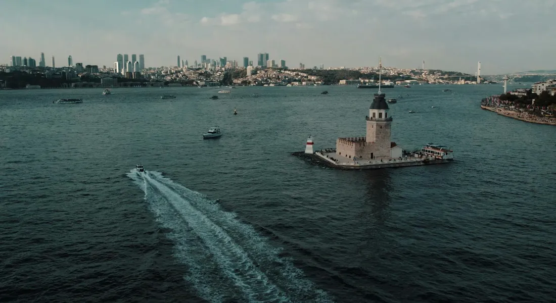Aerial view of a harbor with a small lighthouse on a rocky island, a wake from a speedboat cuts through the water, and a distant city skyline on the horizon.