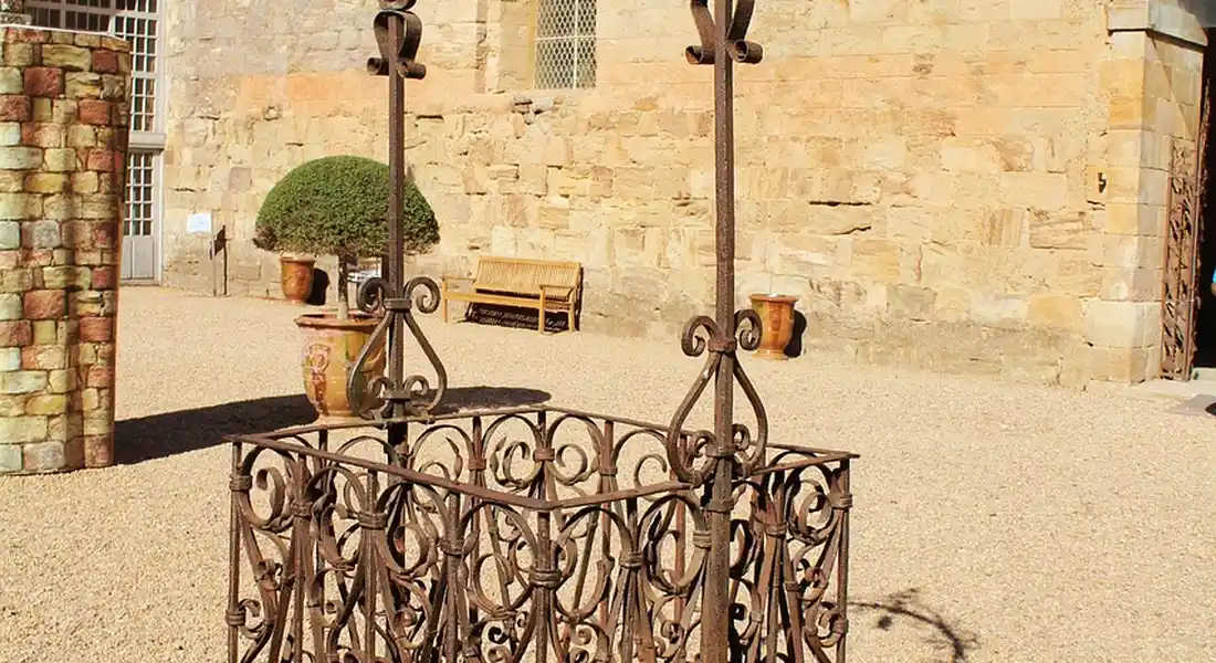 Decorative wrought-iron gate in a sunlit courtyard with a stone wall, potted plants, and a bench in the background.