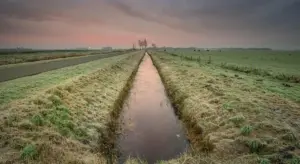 A long, narrow water-filled drainage ditch runs through a grassy field at sunset.
