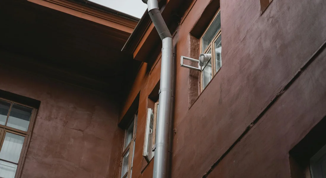 Exterior wall with metal downspout and drainage pipe against a brown building.