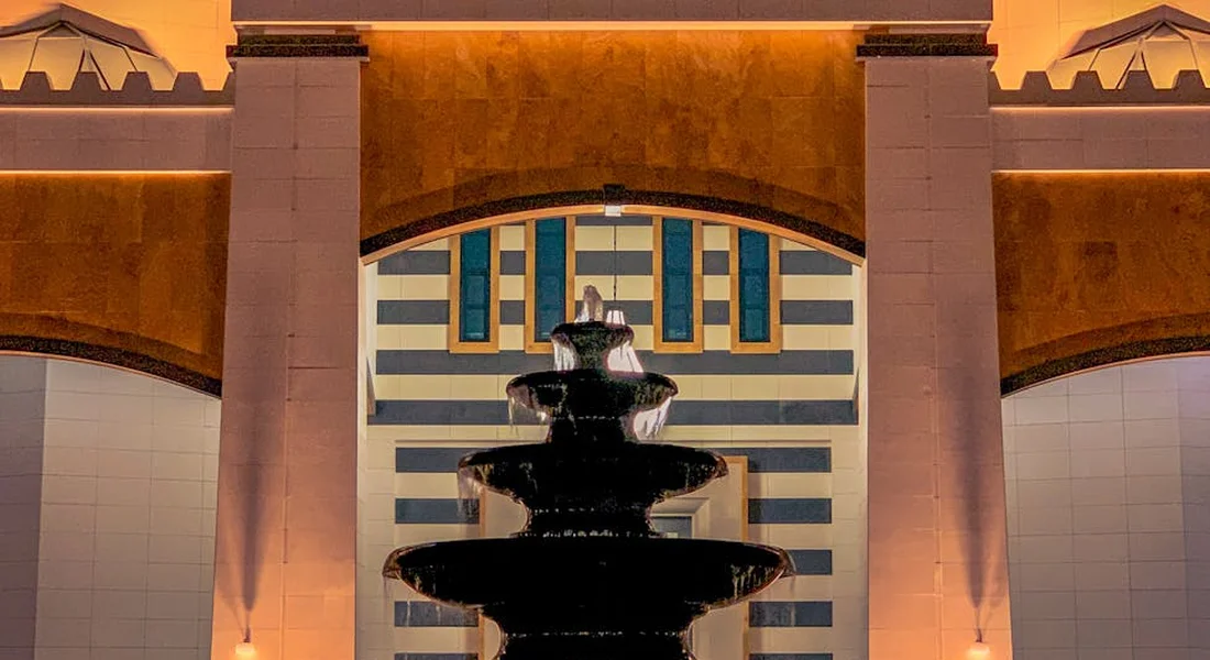 Evening shot of a lit fountain with a modern, arch-shaped building in the background.