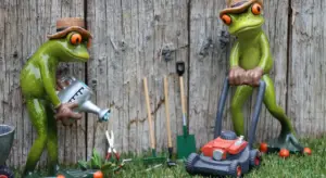 A pair of green frog garden statues wearing hats stand in a yard among garden tools, with a watering can and a toy lawn mower.