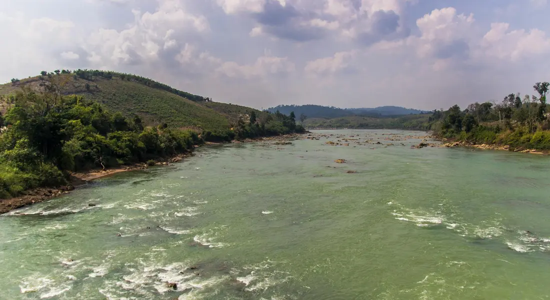 Wide river landscape with greenish water, flanked by hills and a partly cloudy sky.