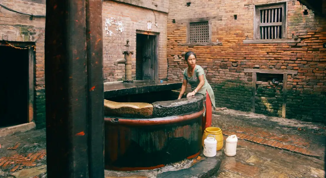 A person standing beside a large round water well in a brick courtyard, with buckets nearby.