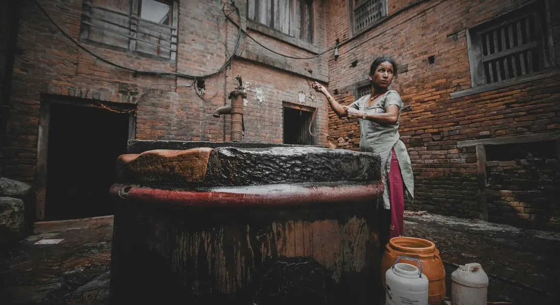 A woman in traditional clothing stands beside a large wooden well in a brick courtyard and operates a hand pump to draw water.