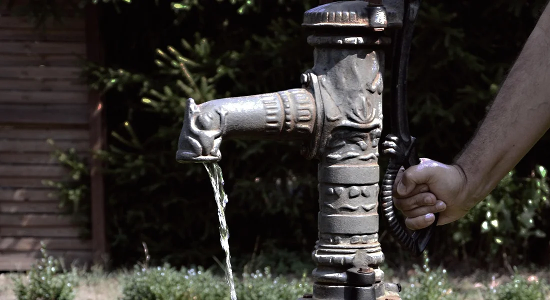 A manual hand pump attached to a domestic water well, with a lever handle and water dripping from the spout.