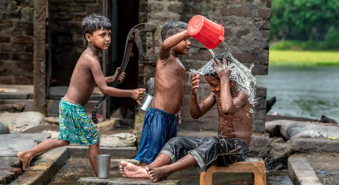Three children by a water source splashing water with buckets, near a brick wall.