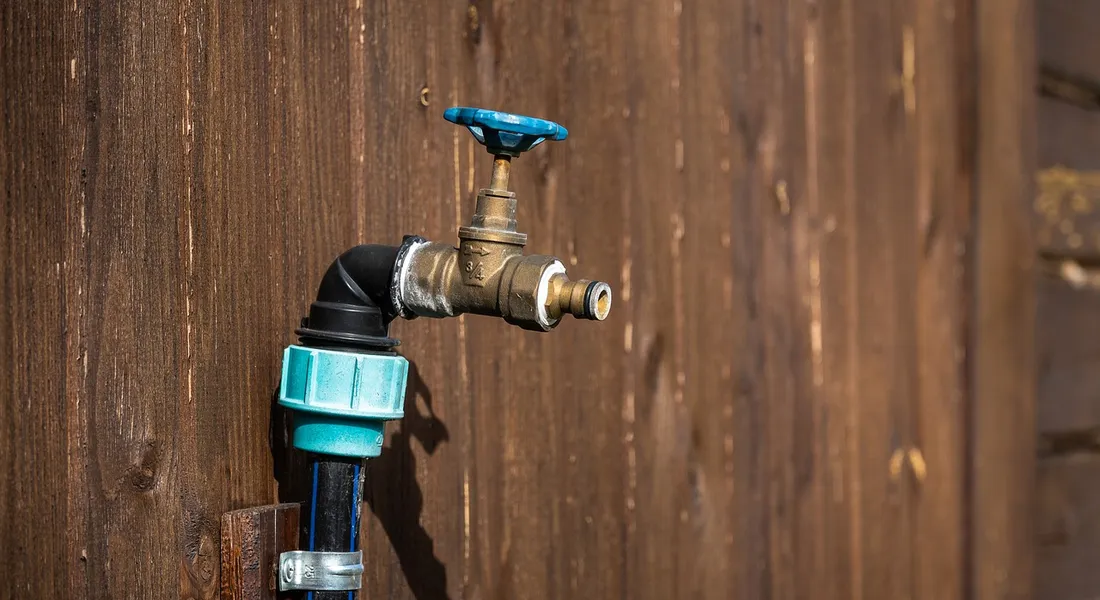 A shutoff valve with a blue handle attached to a water line and a hose connector mounted on a wooden fence, illustrating components used when installing an ice maker water line.