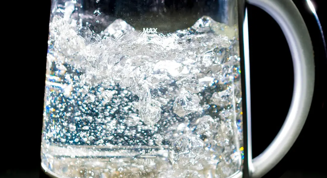 Glass kettle with rapidly boiling water and visible bubbles against a dark background, white handle on the right.
