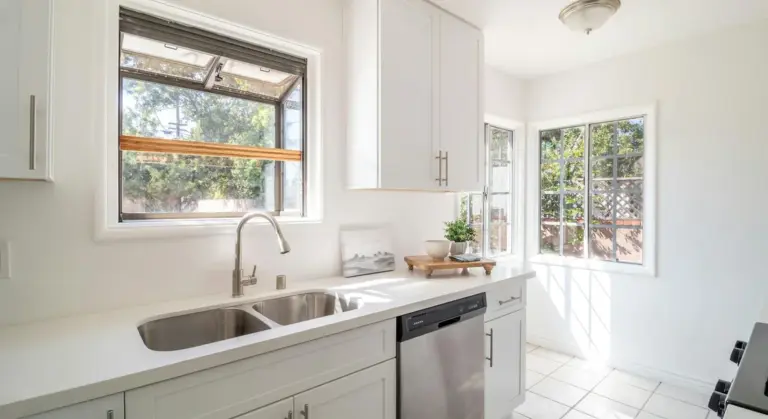 Bright white kitchen with an under-counter stainless steel dishwasher next to a double sink and large windows.
