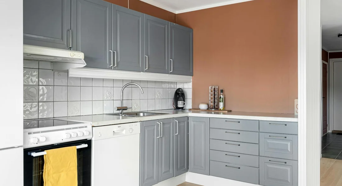 A modern kitchen with gray upper and lower cabinets, a white countertop, and a built-in dishwasher under the counter. An orange accent wall is visible on the right, with a stove on the left and a sink with a faucet in the middle.