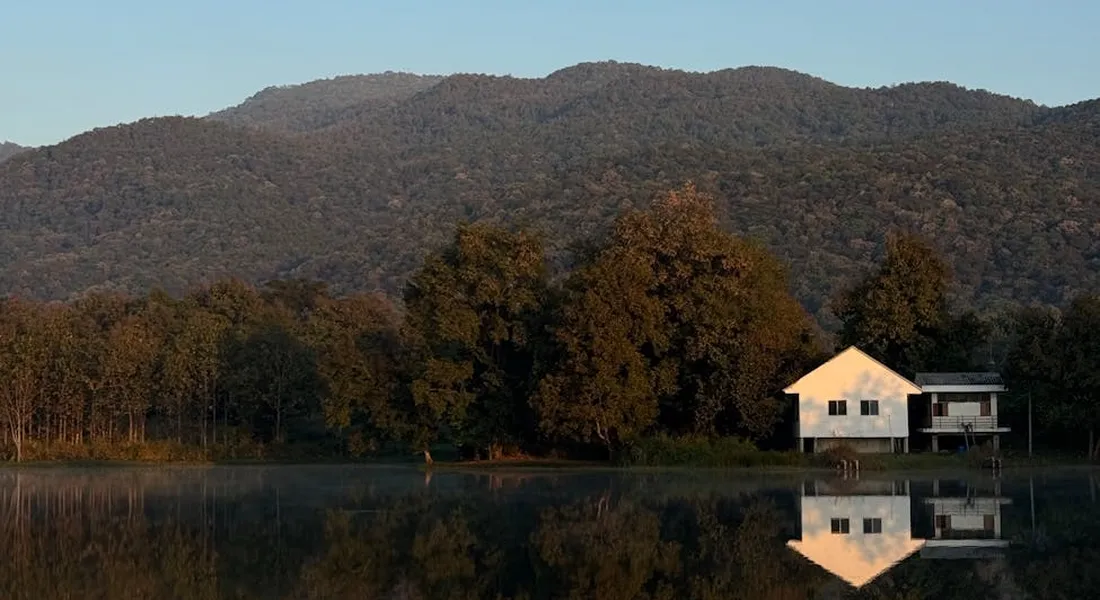 A small white house sits on the edge of a calm lake, with trees and mountains in the background and a clear reflection on the water.