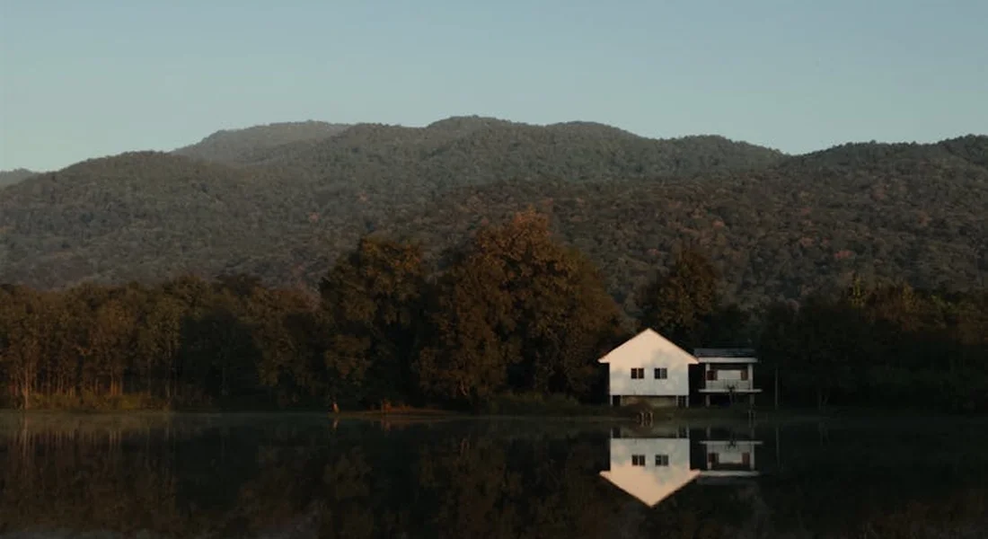 A serene lakeside scene with a white house on stilts reflected in calm water, set against forested mountains.