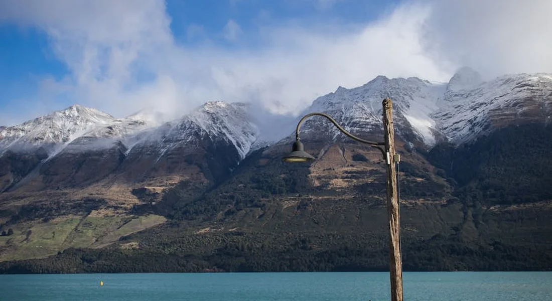 A wooden post with a curved metal lamp over a turquoise lake, with snow-covered mountains and a partly cloudy sky in the background.