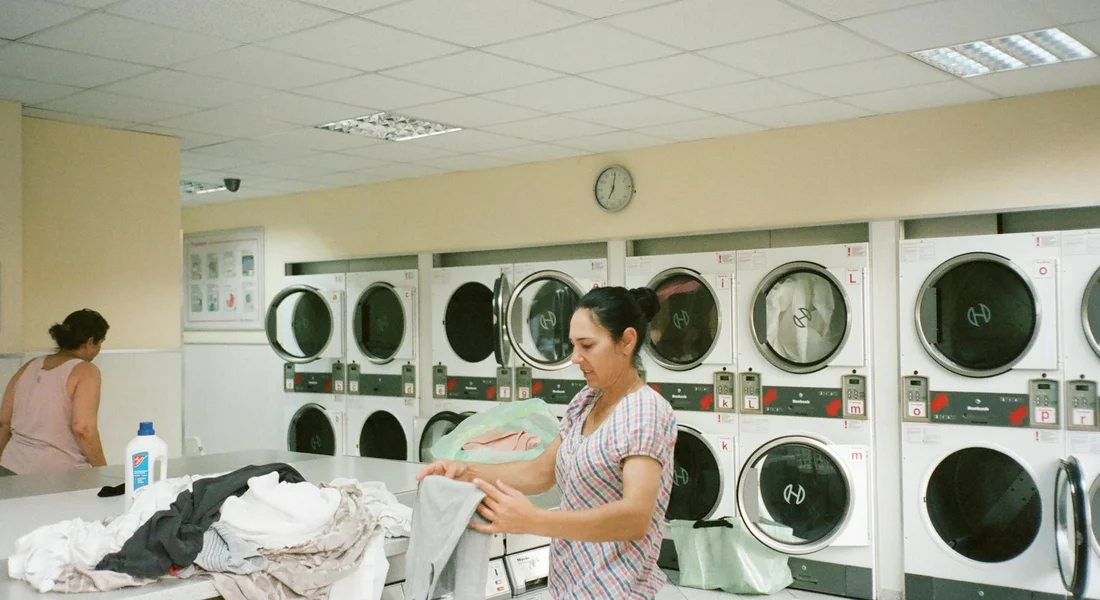 A laundromat with a row of front-loading washing machines; a person folds laundry in the foreground.