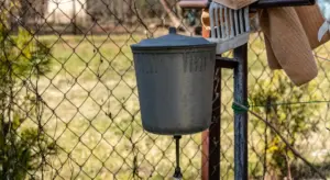 Outdoor metal water storage tank hanging from a post beside a chain-link fence.