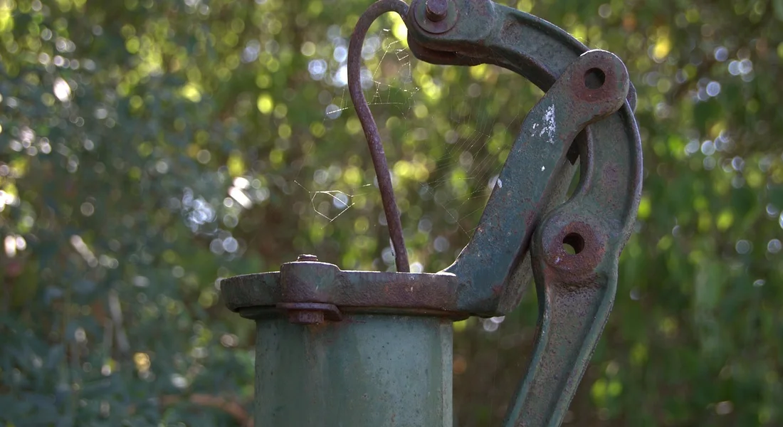 Close-up of a weathered outdoor pump component with a curved metal lever, rusted bolts, and a blurred leafy background.