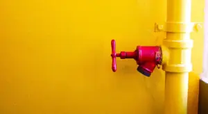 Close-up of a red outdoor water shut-off valve attached to a yellow pipe on a bright yellow wall
