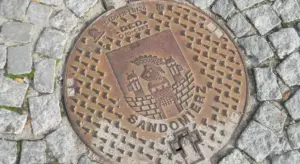 Circular metal manhole cover with a crest emblem on a cobblestone street.