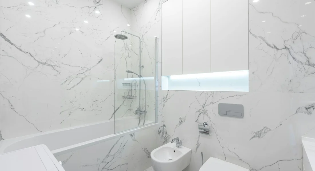 Bright marble bathroom with a wall-mounted toilet featuring a concealed tank, a glass shower enclosure, and a sink