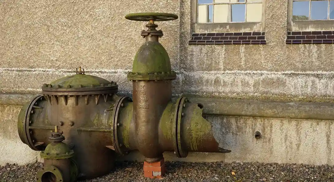 Exterior view of a rusty outdoor water shut-off valve connected to metal pipes against a textured building wall.