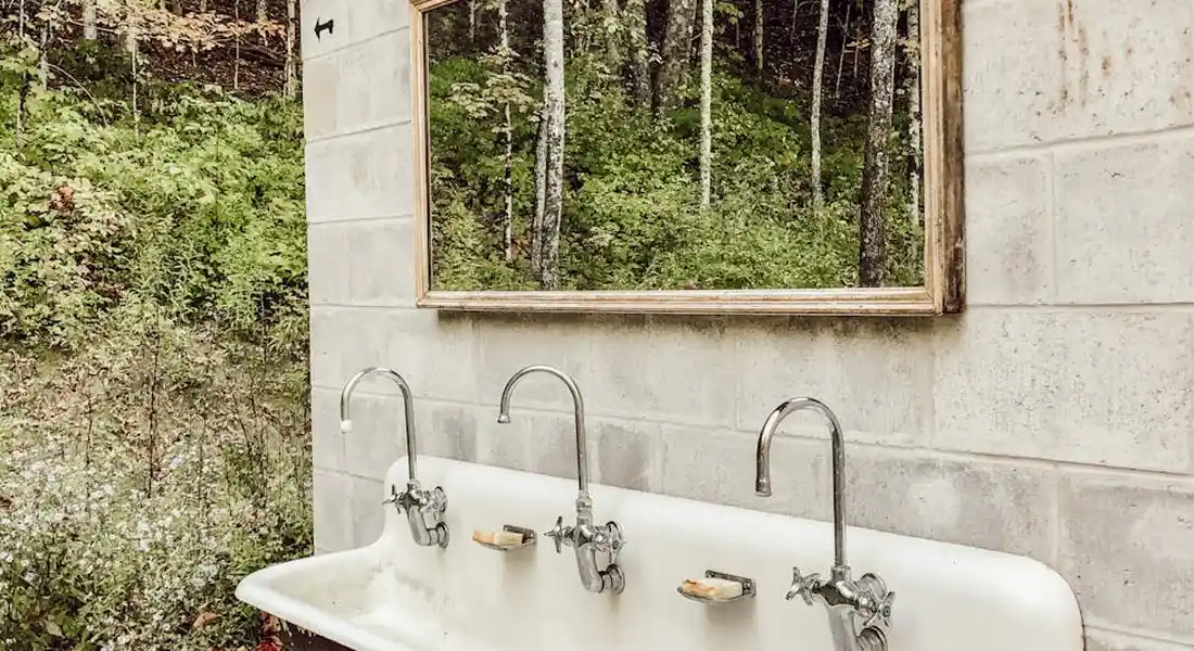 Outdoor utility sink mounted to a concrete block wall with three faucet connections and curved supply lines, set against a forested background.