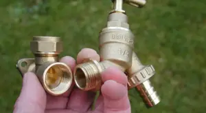 Close-up of a hand holding brass plumbing fittings outdoors, illustrating water service line components.