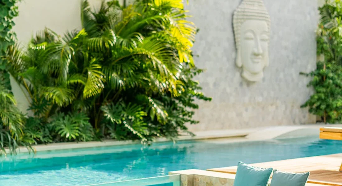 Backyard pool with clear blue water, lush tropical plants along the edge, and a decorative Buddha head wall feature in the background.