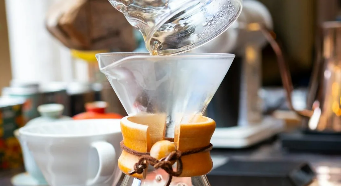 Close-up of a glass pour-over coffee maker with a cone-shaped filter and a wooden collar, set on a kitchen counter with a blurred background.