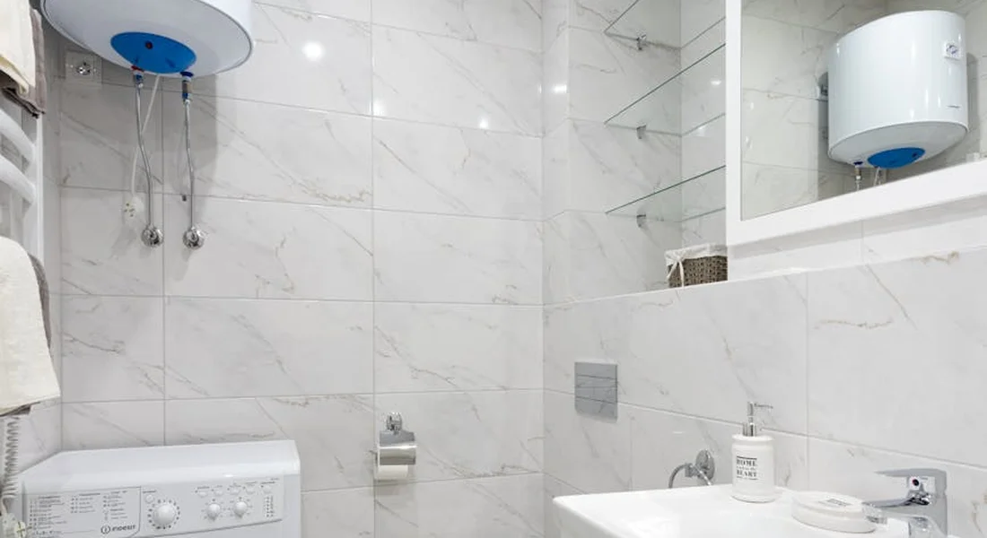 A compact utility bathroom with white tiled walls, featuring a wall-mounted water heater, a washing machine, and a sink.