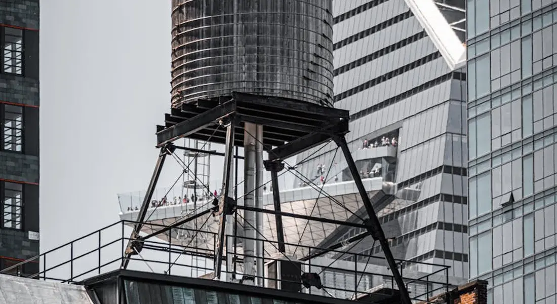 Rooftop water storage tank mounted on a metal frame on a building roof, with piping and a platform, illustrating components for a home water system installation.