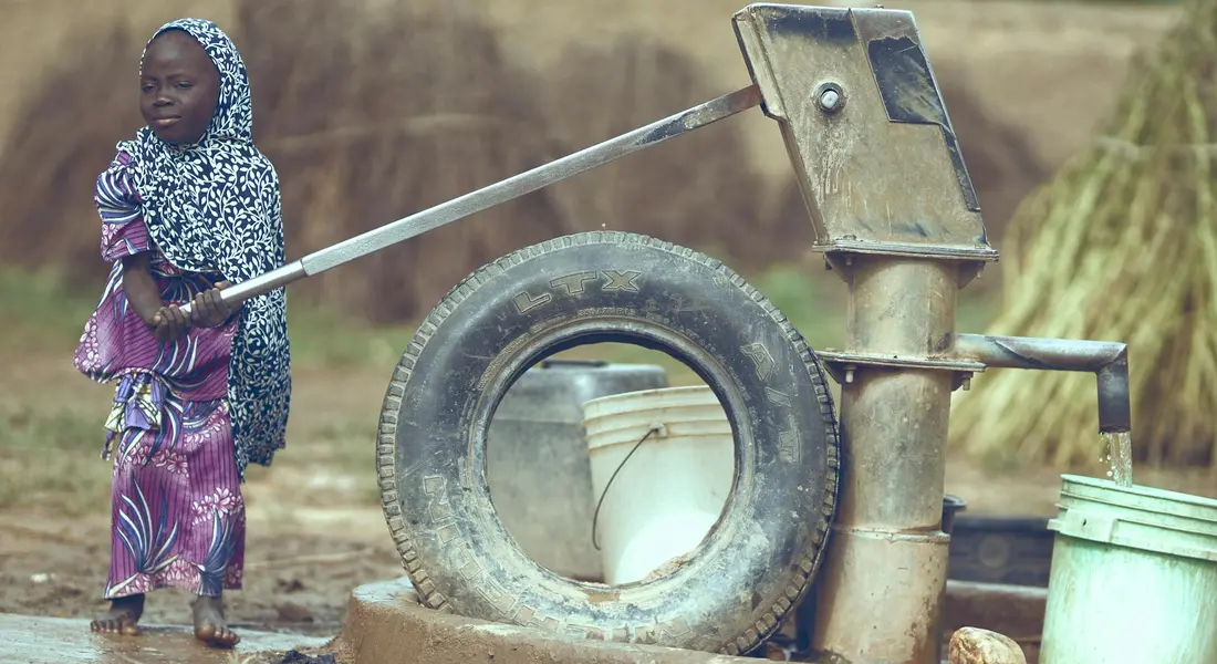 A child standing beside an outdoor hand pump in a rural setting.