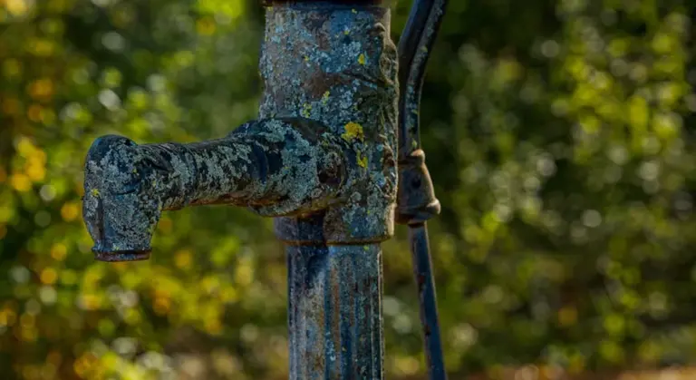 Close-up of a weathered, rusted outdoor water spigot with a blue-green patina, set in a sunlit outdoor scene.