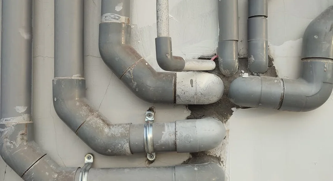 Close-up of a network of gray PVC plumbing pipes and fittings with elbow joints and hose clamps on a wall.