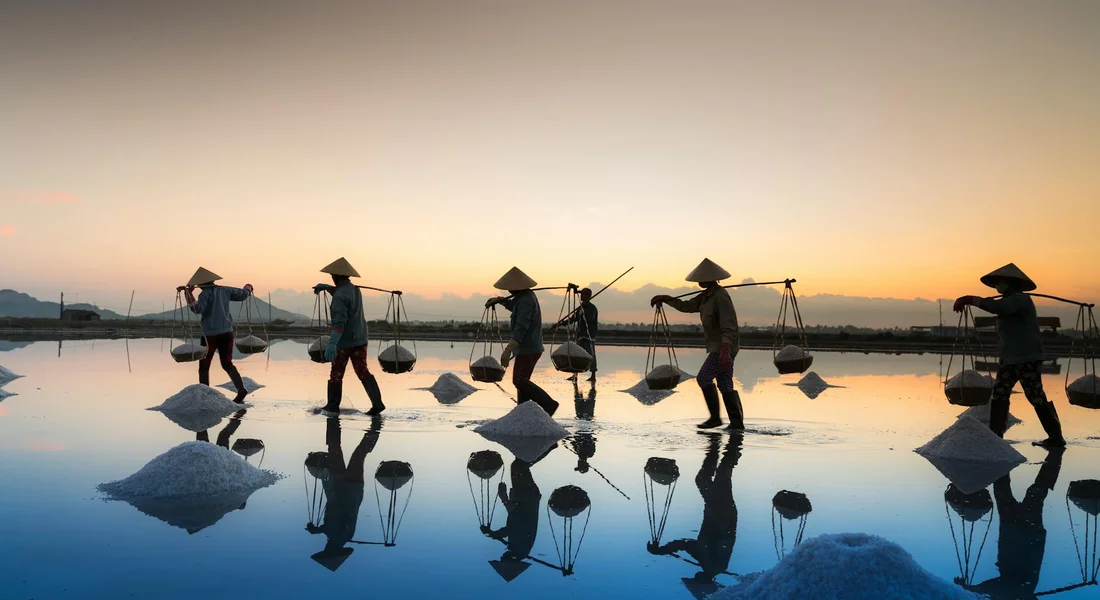 Silhouetted salt workers wearing traditional hats walk across a shallow salt flat at sunset, carrying baskets on poles with piles of salt and reflections in the water.