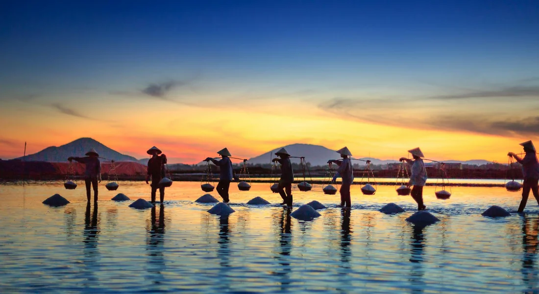 Silhouettes of salt workers in conical hats walking on shallow salt pans at sunset, with reflections on calm water and distant hills.