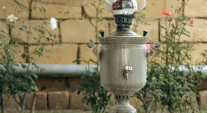 A metal samovar stands on a small outdoor table with plants and a brick wall in the background.