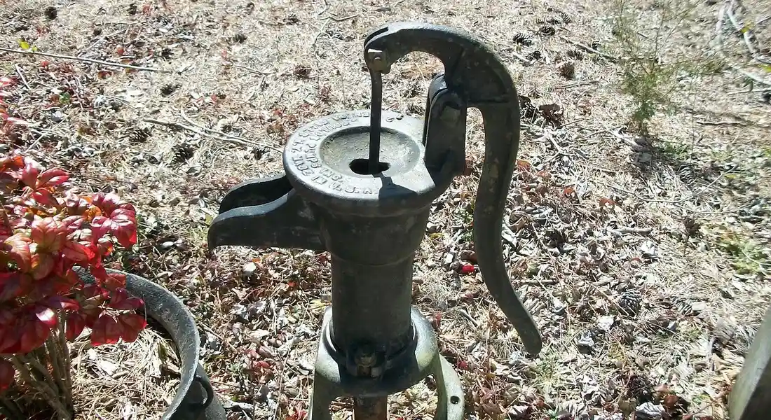 A hand-operated shallow well jet pump mounted outdoors on a stand, with a long lever handle and circular top, surrounded by gravel and plants.