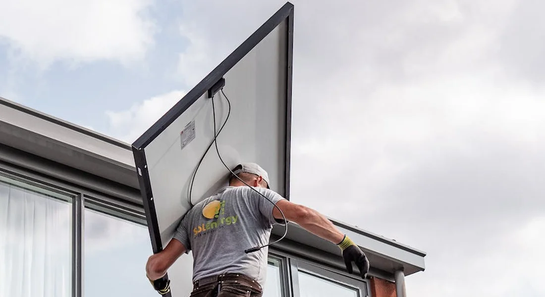 Technician on a roof adjusting a roof-mounted solar water heating panel, wearing gloves and a cap.