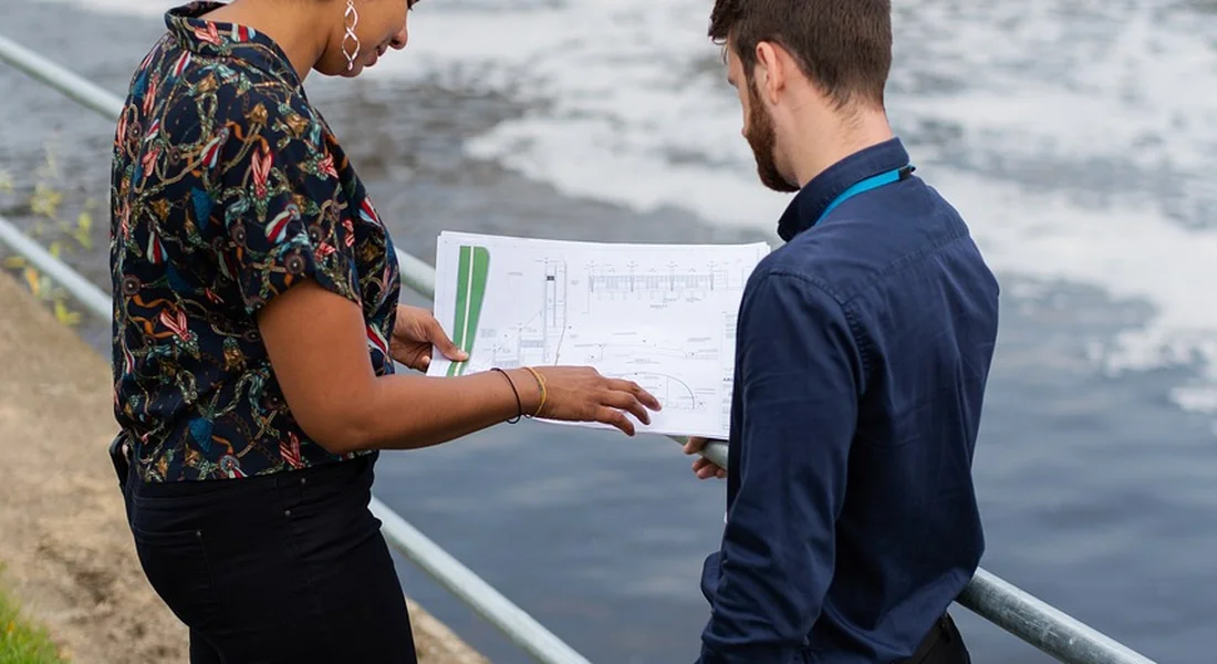 Two professionals review sprinkler system plans near a railing beside a water feature, discussing troubleshooting steps for a backflow preventer.