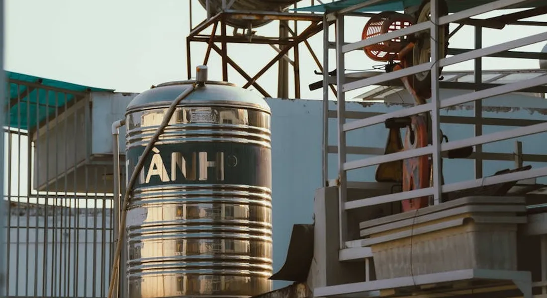 Shiny stainless steel water storage tank on a rooftop with pipes and a staircase in the background