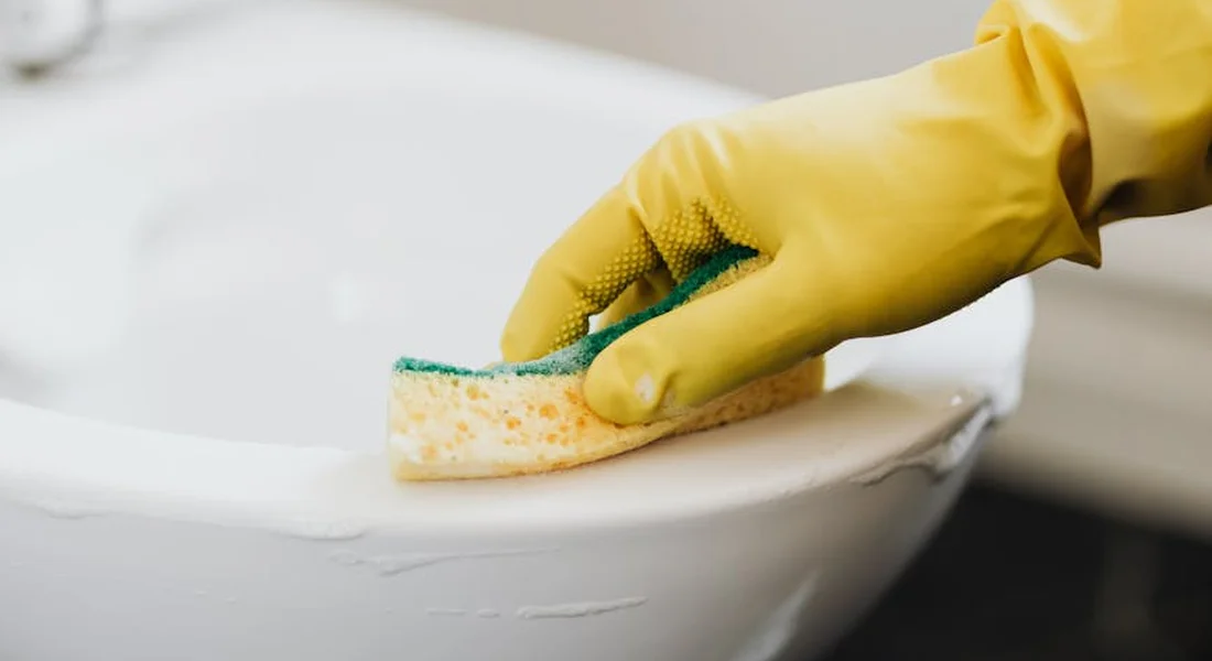 Close-up of a gloved hand using a sponge to clean the rim of a toilet, preparing a dry surface for installing a new water supply line.