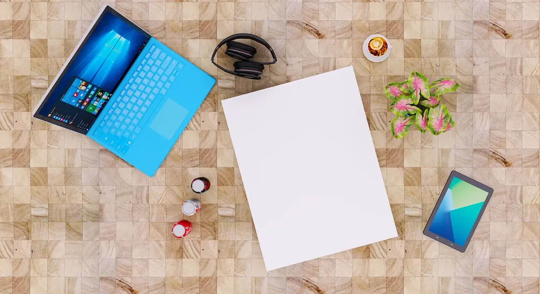 Top-down view of a light wooden desk with a bright blue laptop open, a blank white sheet, a pair of headphones, a tablet, a small plant, and a few tiny bottles or caps scattered nearby.