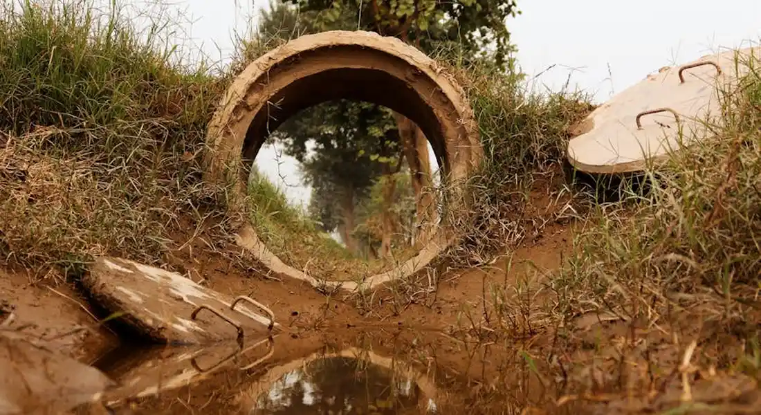 Circular concrete pipe exposed in a muddy trench with wet ground and grass around.